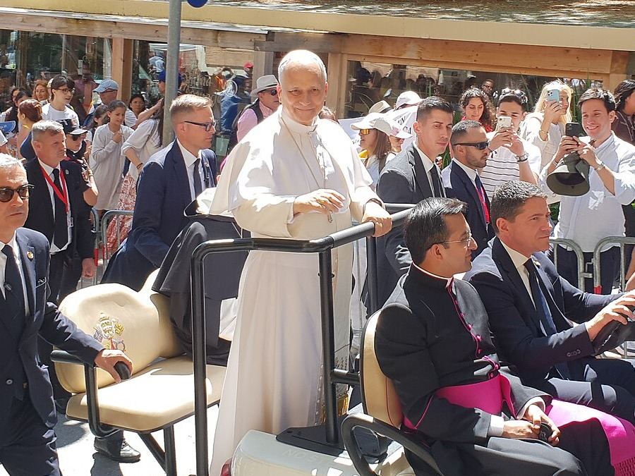 Pope Leo XIV in white, standing in an open cart and greeting a crowd of pilgrims