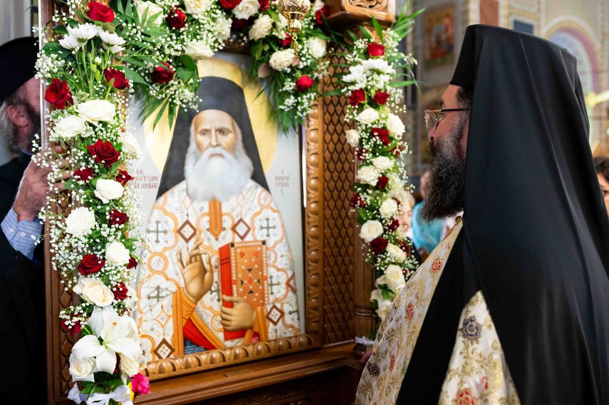 The icon of St. Chrysostomos in a carved frame garlanded with roses and lilies, with a clergyman beside it